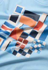 Světle modré bavlněné tričko s barevným geometrickým potiskem obsahujícím čtverce a obdélníky v oranžové, námořnické a modré barvě. Výrazná textura je jemná.