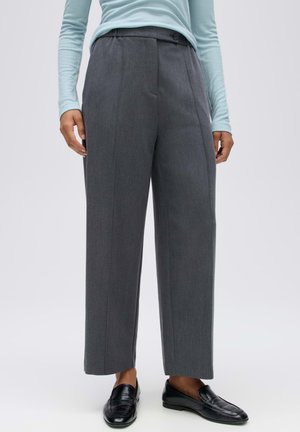 Personne portant un pantalon gris à jambes larges, un haut bleu clair à manches longues et des mocassins en cuir noirs, debout devant un fond uni.