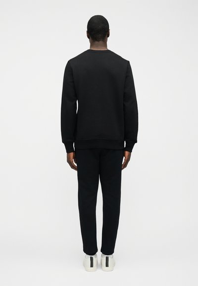 Άντρας που φαίνεται από πίσω, φοράει μαύρο φούτερ και μαύρο παντελόνι με λευκά αθλητικά παπούτσια που έχουν μαύρες και πράσινες ρίγες.