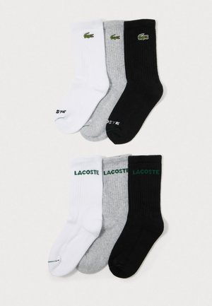 Tři páry sportovních ponožek v bílé, šedé a černé barvě, s logem krokodýla Lacoste a názvem značky na manžetách.