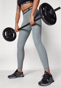 Grå atletiske leggings med tekstureret stof og sort Nike-logo, parret med sorte træningssko med grå accenter. Barbell holdes i hænderne.