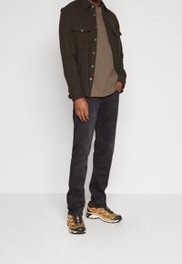 Hombre vestido con una chaqueta abotonada marrón oscuro sobre una camiseta beige, jeans negros y zapatillas de correr naranjas y negras, de pie contra un fondo blanco.