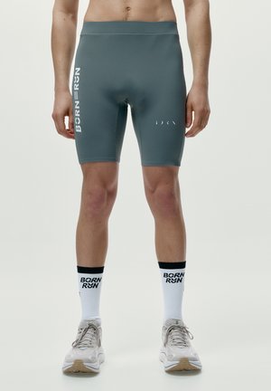 Mann trägt graue BORN RYN Radhosen, weiße BORN RYN Socken und weiße Laufschuhe und steht mit leicht auseinanderstehenden Füßen.
