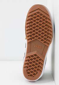 Suela de zapato Vans con una superficie de goma texturizada. Presenta un patrón hexagonal marrón con un logotipo central y un borde blanco.