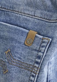 Denimstof in lichtblauw met een gestructureerd oppervlak, voorzien van een leren patch en een messing klinknagel. Zichtbare stiksels in contrasterend bruin.
