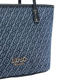 Stor tote-väska i vävd denim med ett marinblått mönster. Har en guldfärgad logotyp och robusta blå läderdetaljer.