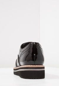 Black patent leather shoe with brogue detailing, rounded toe, and a contrasting beige and black sole, showcasing a smooth, glossy texture.
