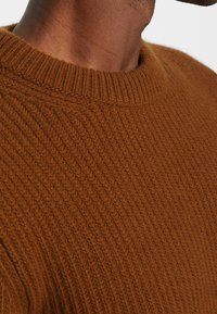 Gros plan sur une personne portant un pull en tricot brun texturé avec un col rond côtelé.