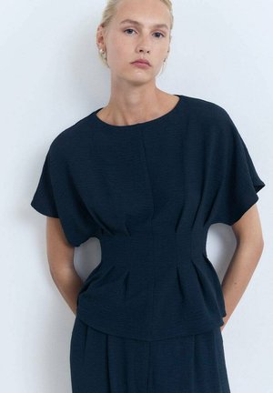 Una modelo con cabello rubio liso que lleva una blusa estructurada de color azul oscuro se encuentra contra un fondo claro, mirando directamente a la cámara.