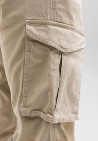 Nahaufnahme einer Tasche mit Klappe an beigefarbenen Cargohosen, die die Stoffstruktur und Nahtdetails an der Seite eines Beins zeigt.