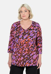 Pitkähihaisen tunikan musta kangas, jossa on eläväinen violetti ja oranssi abstrakti kuvio, syvällä V-aukolla.