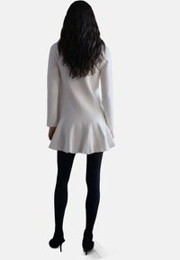 Abito maglione bianco a coste con orlo svasato e plissettato, maniche lunghe, abbinato a collant neri e scarpe con tacco.