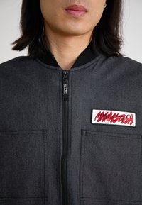 Black bomber jacket made of textured fabric, featuring a front zipper, two chest pockets, and a prominent red embroidered logo patch.