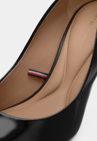 Chaussure plate en cuir verni noir avec une semelle intérieure rembourrée beige et un petit onglet en tissu rouge, blanc et bleu à l'intérieur.