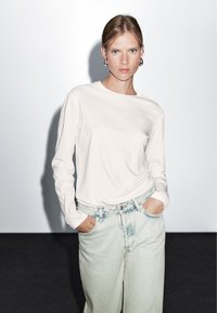 Bílé tričko s dlouhým rukávem a kulatým výstřihem, nošené s džíny s vysokým pasem a světlým ombre. Hladká tkanina, uvolněný střih. Jednoduchý design.