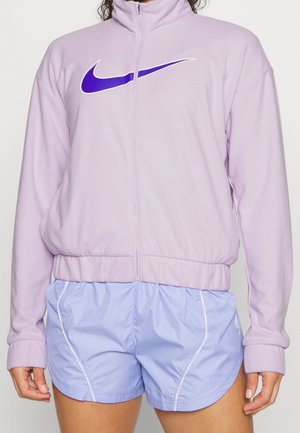 Osoba nosząca jasnofioletową kurtkę zapinaną na zamek z wyraźnym fioletowym logo Nike oraz jasnoniebieskie krótkie spodenki sportowe z białymi szwami.