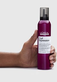 Mousse L'Oréal Curl Expression w ciemnofioletowej butelce z czarną nakrętką. Teksturowana etykieta zawiera szczegóły produktu i składniki. 235 g.
