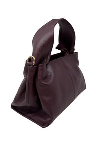 Torebka z ciemnofioletowej, miękko teksturowanej skóry, z szerokim paskiem i metalowymi pierścieniami w kolorze złotym na białym tle.