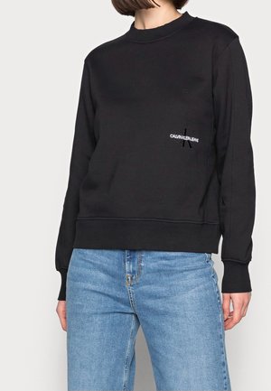 Osoba w czarnej bluzie Calvin Klein Jeans i jasnoniebieskich dżinsach, stojąca na tle jasnego, jednolitego tła, widok od ramion do ud.