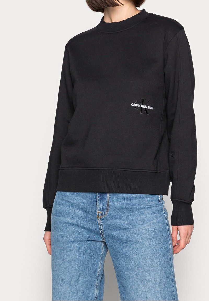 Personne portant un sweat-shirt noir Calvin Klein Jeans et un jean bleu clair, debout devant un fond uni clair, vue des épaules aux cuisses.
