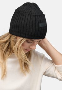 Beanie nera a coste con risvolto, dotata di un'etichetta nera rettangolare. Il beanie ha una superficie testurizzata e una forma aderente.