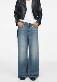 Lichtblauwe wijde jeans met vervaagde wassingen, gecombineerd met een wit T-shirt en een zwart leren kort jasje. Model staat met handen in de zakken.