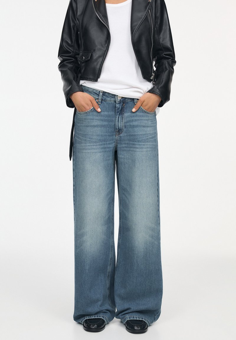 Lichtblauwe wijde jeans met vervaagde wassingen, gecombineerd met een wit T-shirt en een zwart leren kort jasje. Model staat met handen in de zakken.