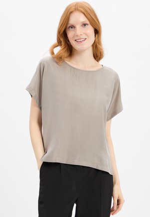 Femme rousse souriante portant un chemisier ample à manches courtes taupe et un pantalon noir, debout avec une main dans la poche devant un fond blanc.