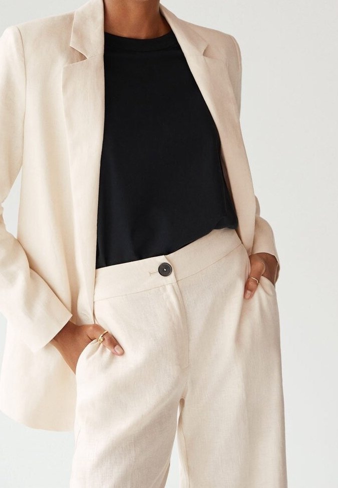 Personne portant un blazer crème et un pantalon assorti, les mains dans les poches sur une chemise noire, montrant des bagues en or aux doigts.