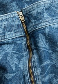 Denimstoff mit blauem Blumenmuster, ausgestattet mit einem goldfarbenen Metallreißverschluss und blauen Nähten entlang der Kanten. Nahaufnahme des Reißverschlussbereichs.