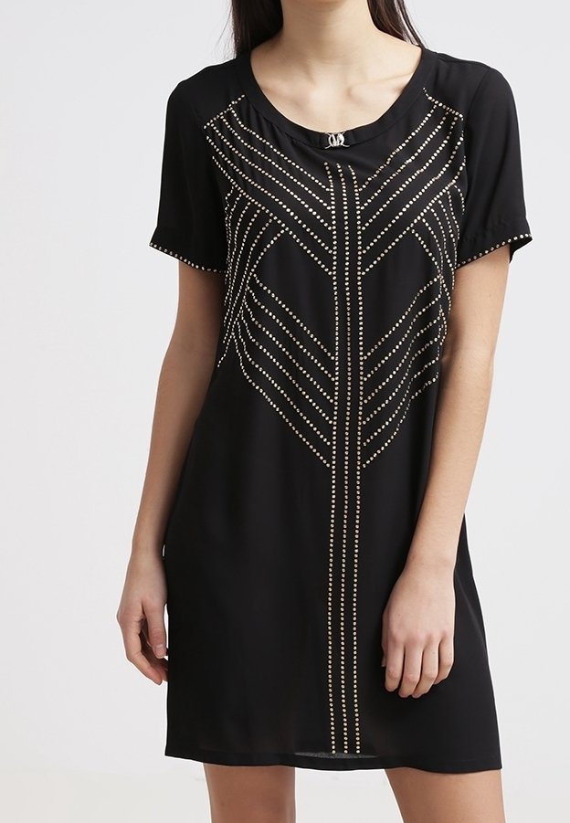 Femme portant une robe noire à manches courtes avec des motifs géométriques cloutés dorés sur le devant et un col rond.