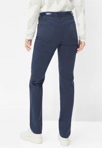 Pantaloni navy blu a gamba dritta con cinque tasche, una texture liscia e una cintura lucida, indossati con sneakers bianche.