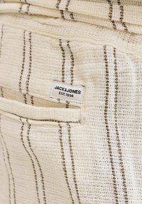 Tissu beige texturé avec des rayures verticales marron et blanches, comportant une petite étiquette blanche portant "JACK & JONES EST. 1990" près du bord d'une poche.