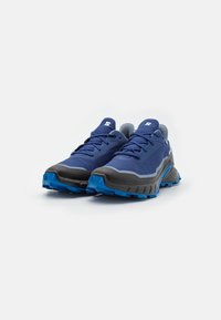 Baskets de sport bleues avec une tige en mesh, accents gris et une semelle noire texturée. Présente un design à lacets et un profil de semelle proéminent pour une meilleure adhérence.