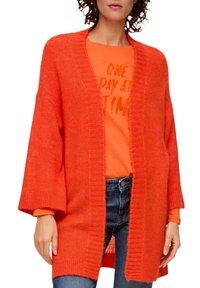 Frau trägt eine übergroße, offene orange Strickjacke über einem orangefarbenen Shirt mit dem Text "EIN TAG NACH DEM ANDEREN" und kombiniert dazu eine blaue Jeans.