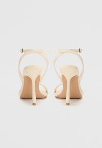 Beige høyhælte sandaler med tynne ankelremmer, åpen tå og en blank overflate. Stiletthæl med en liten gullaksel ved basen.