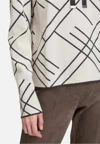 Weißer Strickpullover mit schwarzen geometrischen Linien, verkürztem Design und kontrastierenden Bündchen. Kombiniert mit braunen Wildlederhosen.