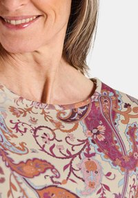 Bluse mit Blumenmuster, die einen cremefarbenen Hintergrund mit Schattierungen von Lila, Pink und Blau zeigt. Weiche Textur und rundem Ausschnitt.