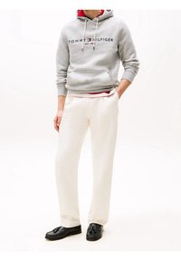 Šedá mikina s kapucí a námořnickým logem, nošená přes červené tričko. Bílé upravené kalhoty a černé polobotky dotvářejí celý outfit.