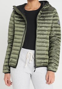 Olijfgroen pufferjack met horizontaal geruit patroon, ritssluiting aan de voorkant, verstelbare capuchon en zwarte voering. Gecombineerd met een zwarte top en grijze broek.