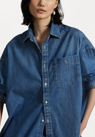 Denimskjorta i mörkblått, med knäppningkrage, framficka och broderad logotyp på bröstet. Mjuk textur med avslappnad passform.