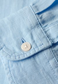 Jasnoniebieska koszula z kołnierzykiem, z białym guzikiem, subtelną fakturą i widocznym ściegiem na materiale.