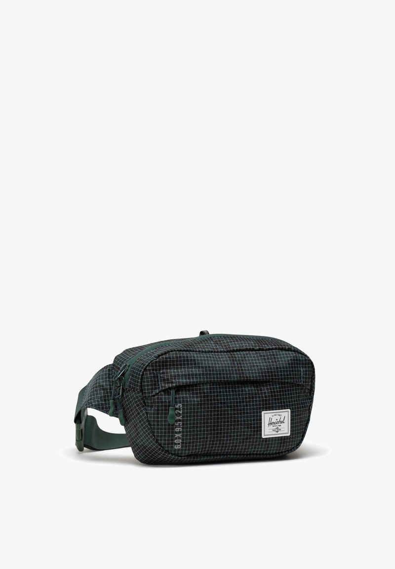 Vihreä ruutukuvioinen kangaskässärilaukku, jossa on vetoketjullinen pääosasto, etutasku ja säädettävä hihna. Sisältää Herschel-brändin logon.