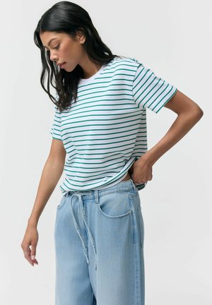 Kobieta poprawiająca jasnoniebieskie spodnie z sznurkiem, ubrana w biały krótki rękaw z zielonymi poziomymi paskami, stojąca na jednolitym tle.