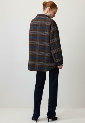 Femme aux cheveux roux attachés en chignon, portant une veste oversize à carreaux bleu et marron, un pantalon noir et des talons foncés, debout devant un fond clair uni.