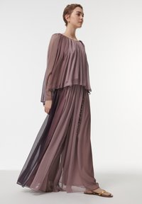 Vloeiende twee-delige outfit in lagen van doorschijnende mauve stof. Bevat lange mouwen en broek tot de enkels met decoratieve knopen langs de zijkant.