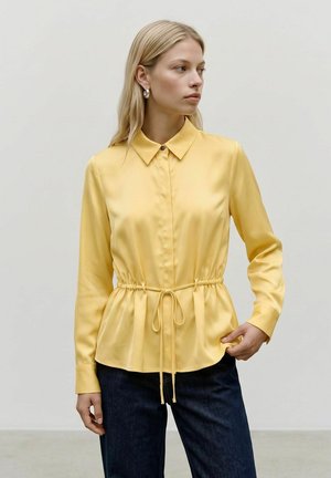 Femme portant un chemisier jaune à manches longues avec col et ceinture à la taille, associé à un jean bleu foncé, debout devant un fond blanc uni.
