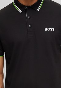 Svart polo-tröja tillverkad av textil med struktur. Har en flerfärgad randig krage, tvåknappars-stängning och en vit broderad "BOSS"-logotyp på bröstet.