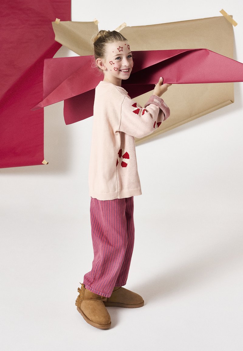 Pull rose en tricot avec des formes d'accent rouges, pantalon rayé rouge et rose, et bottes marron avec des franges ; une fille tient un papier rouge dans un studio.