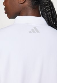 Vrouw met gevlochten haar die een witte textuurpoloshirt draagt met een klein grijs Adidas-logo op de bovenrug.